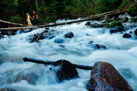 The engaged couple crossing the raging river while balancing on a log during portraits in Frisco, CO