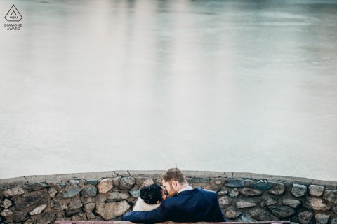 A couple is seated next to a frozen lake, near a stone wall, in Lincoln, Massachusetts, captured beautifully by a talented wedding photographer