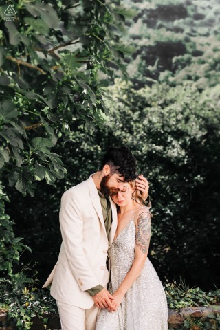 Nel cuore di un giardino di piante nei Paesi Baschi, in Francia, una coppia seduta si tiene per mano su un muro di pietra, condividendo un tenero e caloroso abbraccio prima del loro imminente matrimonio.