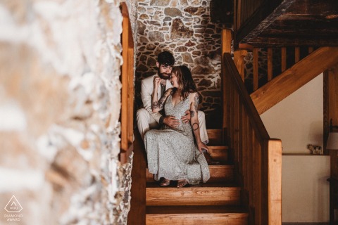 In Basque Country, France, a couple wrapped in love and anticipation sits on the steps of a staircase, as the man envelops the woman in a warm embrace against a large stone wall of an old house, capturing the essence of their upcoming marriage.