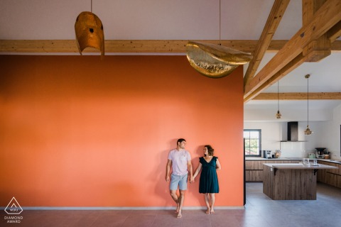 At their engagement portrait session in Basque Country France, the couple stood hand in hand against the large orange wall of their living room, captured in a minimalistic framing that showcased their love and anticipation for their upcoming wedding.