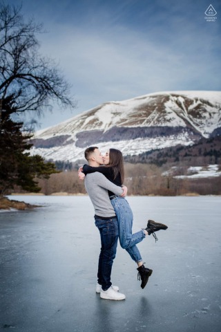During their engagement portrait session at Lac de Petichet, France, the bride and groom-to-be share a playful kiss on the frozen lake, with him lifting her up off the ice in a tender and warm embrace.