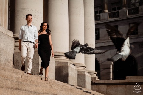 At a London UK prewedding session, a couple of lovers watch as birds take flight on the street nearby, the old world building steps providing a picturesque backdrop near the stone columns.