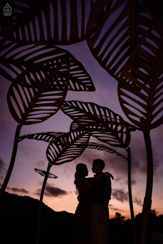 W Samujana w Tajlandii para pozowała do sesji portretowej narzeczeńskiej, a ich sylwetki uchwyciły miłość i oczekiwanie na nadchodzący ślub, ustawione na tle zachodu słońca pod instalacją artystyczną z liści roślin na zewnątrz.