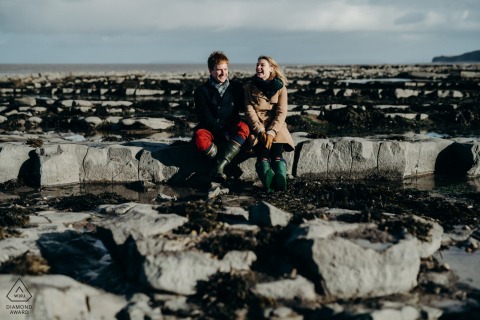 At Porlock Beach, a couple sits together on the rocks, laughing and dressed warmly for their engagement portrait session before their upcoming wedding.
