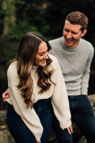 Im Botanischen Garten in Bath beugt sich das Mädchen vor und lächelt mit ihrem Mann, während sie während ihrer Verlobungsporträtsitzung über seinen Witz lacht.