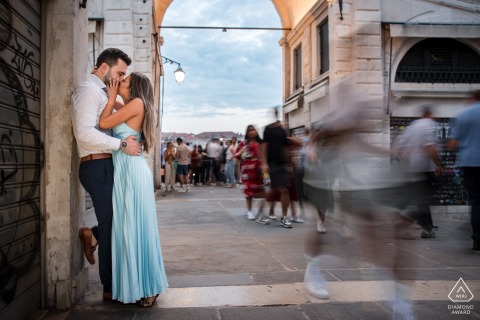 At the Rialto Bridge in Venice, a romantic couple in love, who are engaged to be married, share a kiss while passersby walk past them, in a blur of motion.