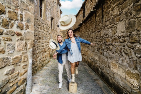In Baeza, Jaén, Spain, before their wedding, a couple poses playfully together, throwing their hats towards the camera in a charming alleyway with ancient stone walls.
