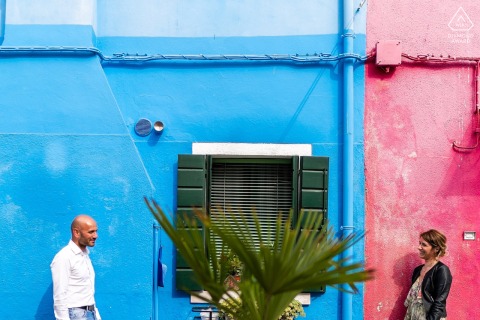At Burano, Venezia, Italy, a couple in front of a double-colored wall of pink and blue, stand opposite each other, gazing into each other's eyes during their engagement portrait session.