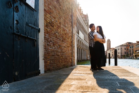 At Venice, Italy, a couple hugged near the Canal Grande, standing by historic ancient-looking docks and walls of stone to take photos before their wedding.