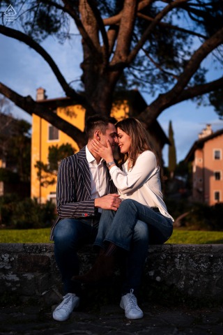 Em Gorizia, Itália, um casal estava sentado num banco, abraçando-se sob uma grande árvore durante o pôr do sol, com uma luz quente iluminando lindamente a cena.