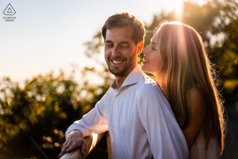 In Duino, Trieste, Italy, a couple is hugging during the beautiful golden hour, with her arms wrapped around him and both of them smiling with their eyes closed.