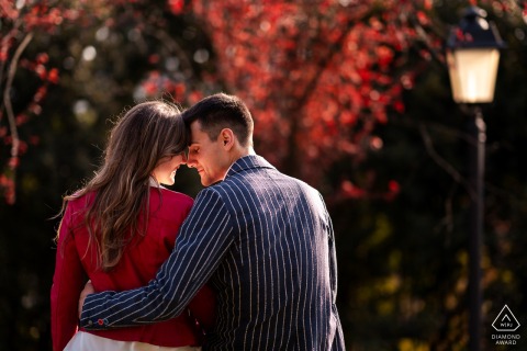 In Gorizia, Italy, a couple sits on a wall hugging each other in a cozy setting with red leaves on trees in the background for their engagement photo shoot.