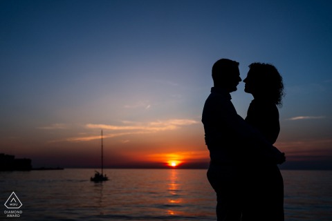 In Trieste, Italy, a couple stands in silhouette against the colorful sunset with a sailboat in the background, capturing their love and excitement during their engagement portrait session.