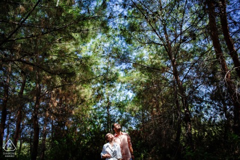 During a photo session at Korineum Hotel in Kyrenia, Cyprus, the couple poses in a symmetrical shot with trees surrounding them, looking up towards the sky from a low position in the frame.