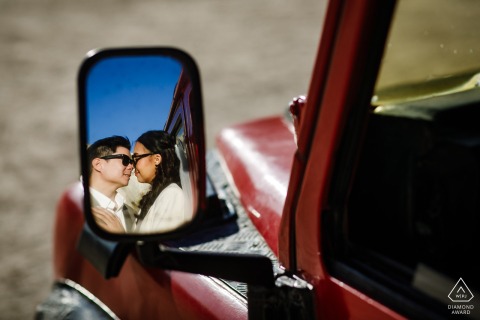 In Cappadocia, Turkey, a couple preparing for marriage poses for photos wearing sunglasses while looking at each other, their reflection seen in the side mirror of a classic red car.