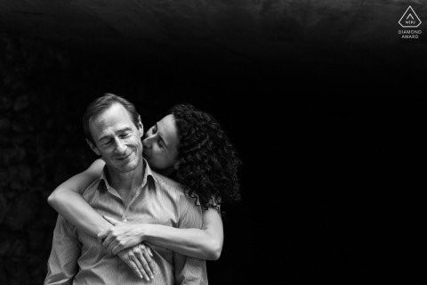 In a medieval village called Pérouges in France, a couple getting married posed for a black and white photo. The woman hugs her partner from behind, kissing his neck while he smiles with closed eyes.