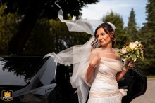 In Gorizia, Italië, stapt een bruid op haar trouwdag gracieus uit een zwarte auto, haar sluier elegant gevangen door de wind, wat een vleugje verfijning en stijl toevoegt aan dit glamoureuze moment.
