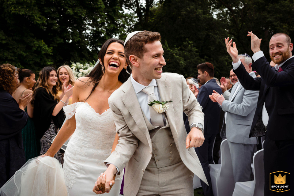 At Sopwell House in the UK, the bride and groom walk down the aisle with beaming smiles outside, surrounded by friends, creating a heartwarming scene of love and companionship.